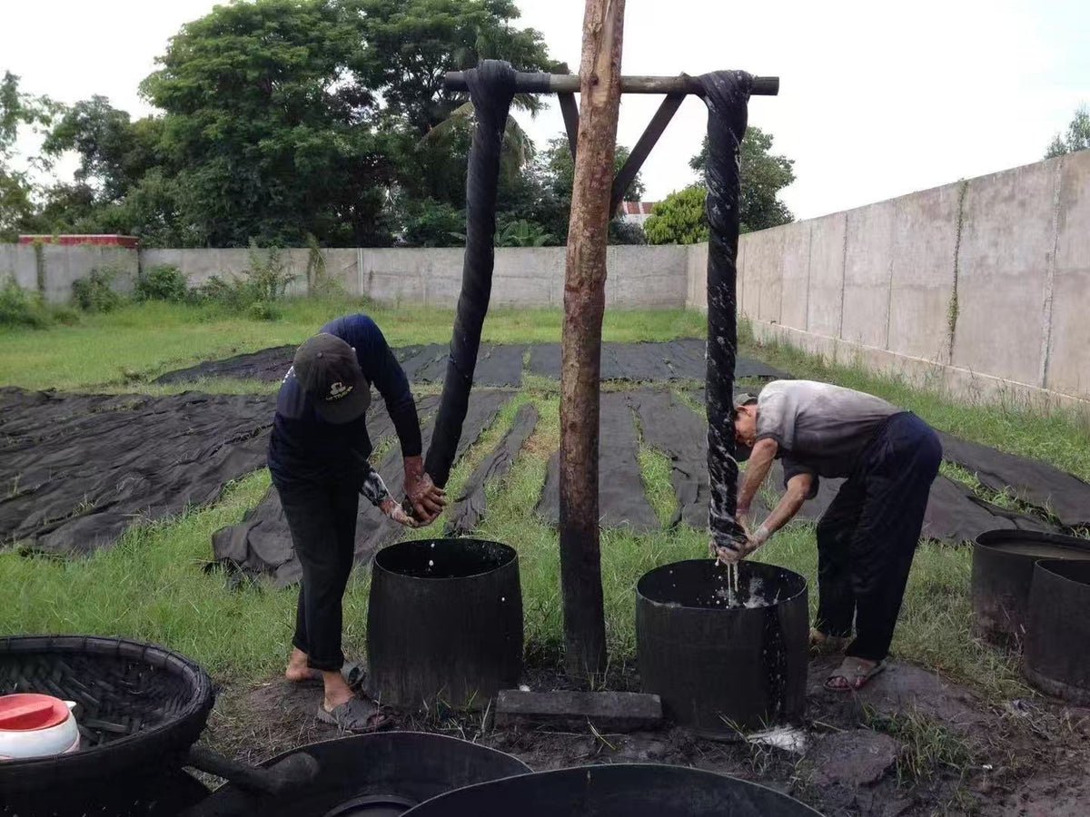 Craftspeople wringing naturally dyed fabric by hand on a wooden frame, with lengths of fabric drying on the grass